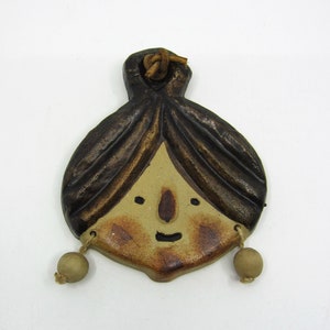 Op de afbeelding: Een keramische ornament met een afbeelding van een vrouwen gezicht met bruin haar, bruine ogen en een kleine neus. De ornament heeft een bruin leren koord en twee kleine beige kralen die aan de onderkant hangen.