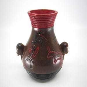 Puede incluir: Un jarrón de cerámica marrón con un borde rojo y dos asas con forma de cabezas humanas. El jarrón presenta figuras grabadas en rojo y negro, incluyendo formas humanas y animales. El jarrón tiene un cuerpo bulboso y un cuello acanalado.