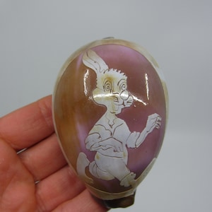 Puede incluir: Una concha de mar rosa y marrón con una imagen blanca tallada de un conejo que lleva una camisa y pantalones.