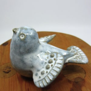 Può includere: Figurina di uccello in ceramica nei toni del blu e del bianco, con dettagli sulle ali e sulla coda. L'uccello è appoggiato su una superficie di legno. La figurina ha un corpo arrotondato e un piccolo becco.