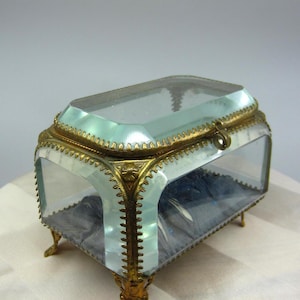 Peut inclure: Une petite boîte à bijoux en verre transparent avec un cadre et des pieds en métal doré. La boîte a un couvercle à charnière et une doublure en velours.