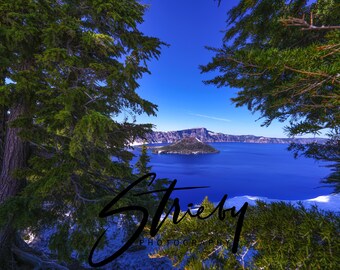 May include: A scenic landscape photograph featuring a deep blue lake surrounded by mountains and evergreen trees. The sky is a clear, bright blue. The image is framed by green foliage, with the word "Strieby PHOTOGRAPHY" in the foreground.
