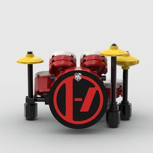 May include: A miniature drum set made of red, black, and yellow blocks. The bass drum features a black face with a red logo. Two cymbals are positioned above the drums. The set is on a white background.