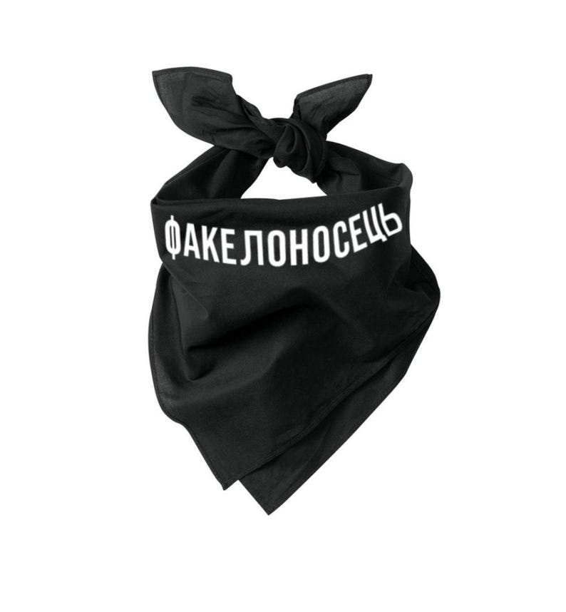 May include: Black bandana with white text reading "ФАКЕЛОНОСЕЦЬ". The bandana is tied at the top, creating a knot. The fabric is folded at the bottom. The bandana is made of a soft material.