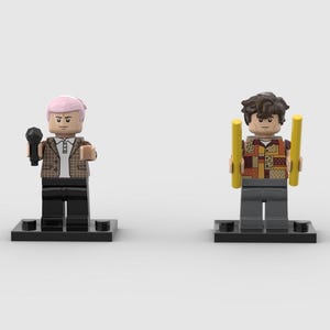May include: Two Lego figures on black bases. One figure has pink hair, a brown plaid jacket, and holds a black microphone. The other figure has brown hair, a patchwork shirt, and holds two yellow batons.