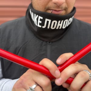 May include: A black neck gaiter with white text that reads "KEЛOHOP" is worn over a gray long-sleeve shirt and black vest. Two bright red poles are held in front of the person. Silver rings are on the person's fingers.