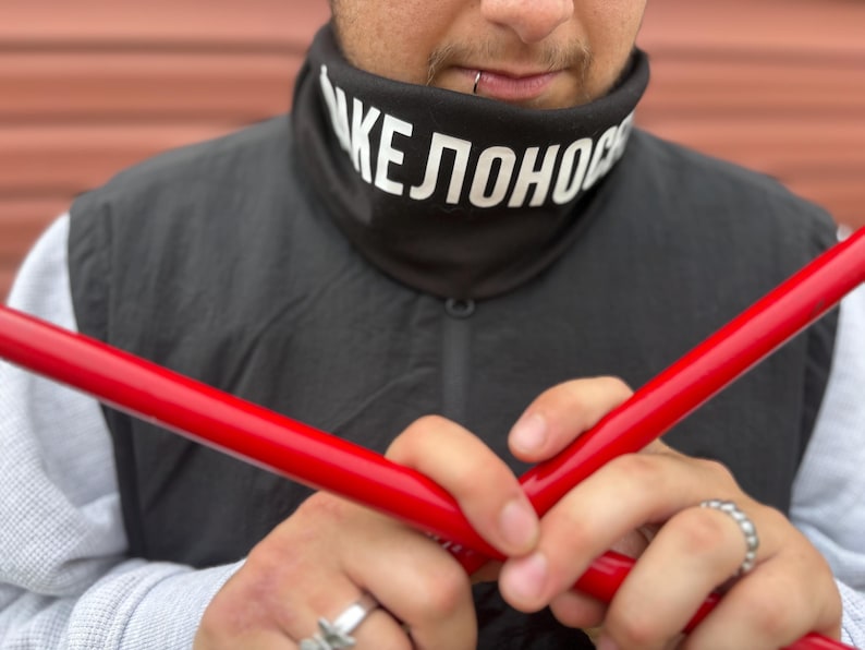 May include: A black neck gaiter with white text that reads "KEЛOHOP" is worn over a gray long-sleeve shirt and black vest. Two bright red poles are held in front of the person. Silver rings are on the person's fingers.