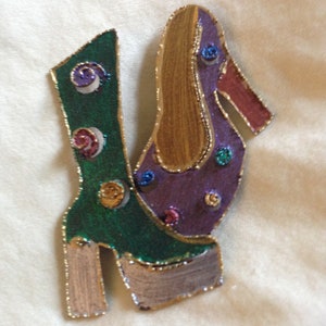 May include: A whimsical, handcrafted brooch in the shape of a high-heeled boot. The brooch features a green boot with gold trim and colorful accents. The shoe's heel and platform are painted in a metallic gold and silver. The design is embellished with small, colorful circular accents.