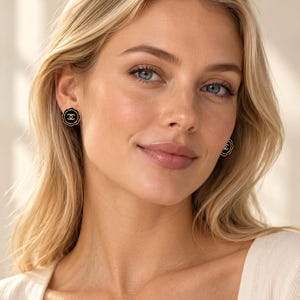 May include: A pair of round stud earrings featuring a black circular center with a gold-toned interlocking CC logo. The earrings have a scalloped gold-toned edge, creating a flower-like silhouette that sits flush against the earlobe.