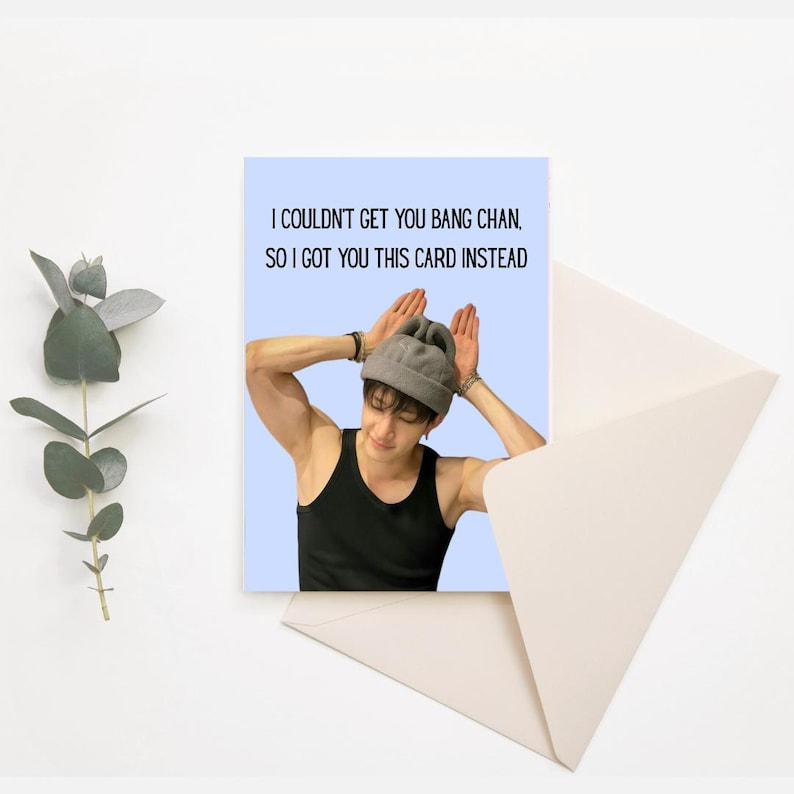 May include: A greeting card featuring a photograph of a man wearing a grey beanie and making bunny ears with his hands. The text on the card reads "I couldn't get you Bang Chan, so I got you this card instead."