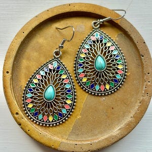 May include: Silver-coloured teardrop earrings with colourful geometric patterns. Each earring has a turquoise teardrop-shaped stone at the centre, surrounded by a filigree design and a border of small, colourful diamonds. The earrings are displayed on a round, textured, tan-coloured surface.
