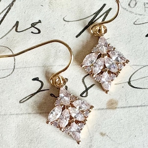May include: A pair of gold earrings featuring a diamond-shaped cluster of clear, marquise-cut gemstones. The earrings have a delicate hook design, set against a vintage paper background.