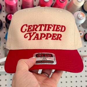 May include: A cream-colored baseball cap with a red brim. The cap features the embroidered text "CERTIFIED YAPPER" in red. The hat is held up in front of a wall of colorful spools of thread.