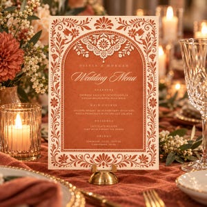 Könnte beinhalten: Eine rechteckige Hochzeitsmenükarte mit terrakottafarbenem Hintergrund und weißem Text. Der Rand zeigt komplizierte florale und vogelartige Motive in Weiß. Der Text lautet Olivia & Morgan Wedding Menu und listet Vorspeisen, Hauptgerichte, Desserts und Getränke auf.