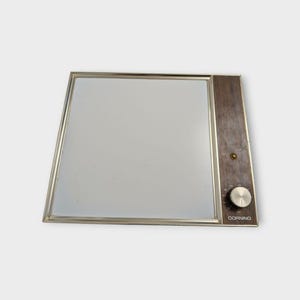 May include: A square electric warming tray with a white glass surface and a metallic silver frame. The right side features a dark wood-grain panel with a small circular indicator light, a silver control knob, and the word CORNING printed in white.