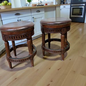 Könnte beinhalten: Zwei Barhocker aus braunem Leder mit dunklen Holzrahmen. Die Hocker haben runde, gepolsterte Sitze und dekorative Schnitzereien an den Beinen und Zargen. Die Sitze sind in einem satten Braunton gehalten, und das Holz hat einen warmen, rötlich-braunen Farbton.