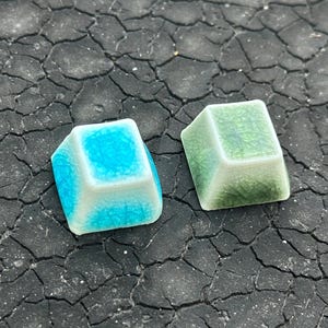 Pode incluir: Dois objetos de cerâmica em forma de cubo. Um é turquesa e branco, o outro verde. Ambos têm um acabamento craquelado. Os cubos estão sobre uma superfície escura e texturizada.