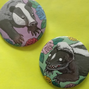 May include: Two round button pins featuring illustrations of forest animals. The first pin features a badger with a pink and purple floral background. The second pin features a skunk with a green and yellow floral background. The text on the card reads "Forest Friends, A button set by Tentacledoom, shoppeofdoom.etsy.com".