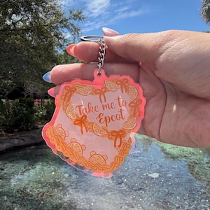 May include: A pink and orange keychain with the text "Take me to Epcot" in a decorative font. The keychain has a decorative border and a silver chain. The background shows a sunny sky and a water feature.
