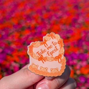 May include: A heart-shaped orange and pink enamel pin with the text "Take me to Epcot" on a white background. The pin is in the shape of a cake with orange slices and bows.