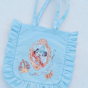 May include: Pale blue tote bag with frilled edges and matching handles. The bag features a framed illustration of Mickey Mouse in a sorcerer's outfit, with the word "FANTASIA" and other graphics. The bag is made of a smooth, possibly nylon, material.