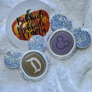 May include: Two white glitter mouse ear keychains with a purple and gold button. The purple button has a Mickey Mouse silhouette and the gold button has a Disney "D" logo.
