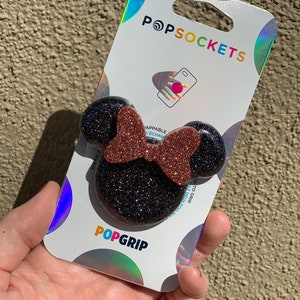 May include: A black and brown glitter PopSocket phone grip with a Minnie Mouse design. The PopSocket is on a white background with the word "PopSockets" printed in black.