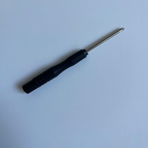 May include: A black screwdriver with a silver metal tip. The handle is black and has a textured grip. The screwdriver is lying on a white surface.