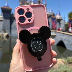 May include: A light pink smartphone case featuring a triple-lens camera cutout and a black, balloon-shaped grip attachment on the back. The grip displays a small, white, outlined Mickey Mouse head icon centered within the black balloon silhouette.