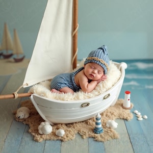 May include: A newborn baby wearing a blue and white striped hat and romper sleeps peacefully inside a white toy sailboat. The boat rests on a bed of faux fur and sand, with miniature lighthouses and seashells nearby. The background features a painted ocean scene.
