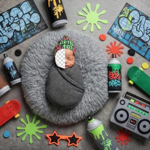 May include: A baby wrapped in a grey blanket, wearing a colourful beanie, surrounded by graffiti-themed props. Includes toy spray paint cans, skateboards, and a boombox, with colourful paint splatters and graffiti art.