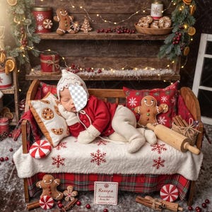 May include: A baby dressed in a red Christmas outfit and a white hat is lying on a miniature wooden bed. The bed is decorated with gingerbread man pillows, a rolling pin, and a blanket with snowflake and gingerbread man designs. The scene is festive with Christmas decorations.
