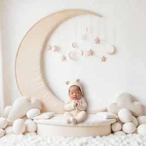 May include: A newborn baby in a beige bear hat and jumpsuit sits on a round platform. Cloud and oval-shaped pillows surround the platform. A large beige crescent moon backdrop with hanging stars and moons completes the nursery scene.