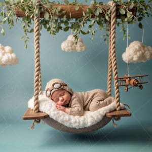 May include: A baby wearing a brown aviator hat and goggles sleeps in a white and brown swing. The swing is suspended from a wooden branch with rope. There are white fluffy clouds hanging from the branch above the swing. A small wooden airplane hangs from the branch on the right side of the image.