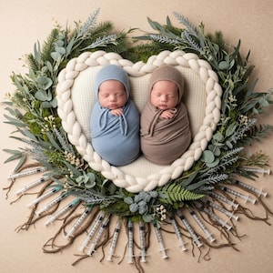 May include: Two newborn babies wrapped in blue and brown swaddling blankets and matching caps, resting inside a heart-shaped wreath. The wreath is made of greenery and a braided white fabric. Syringes are arranged around the base.