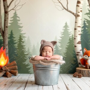 May include: A newborn baby wearing a brown knitted hat is sleeping in a silver bucket. The baby is surrounded by a woodland backdrop with trees, a campfire, and a fox.