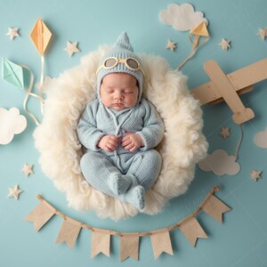 May include: A baby in a light blue aviator outfit, resting in a fluffy white nest. The image features paper airplanes, clouds, stars, and a banner against a light blue backdrop, creating a whimsical scene.
