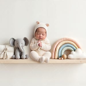 May include: A newborn baby wearing a white knitted outfit with bear ears, sitting on a wooden shelf with a gray elephant plush toy, a rainbow-colored arch, and a white cloud plush toy.