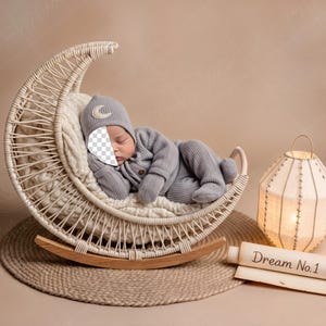 May include: A newborn baby in a gray outfit and hat with a crescent moon design, sleeping in a woven crescent moon-shaped cradle. A decorative lantern and a sign that reads "Dream No. 1" are also in the image.