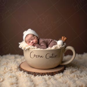 Puede incluir: Un bebé recién nacido dormido, con gorro blanco y traje marrón, dentro de una taza beige. La taza, con "Cocoa Cutie" escrito, está sobre una rodaja de madera. Una rama de canela y una nube de azúcar decoran el borde. Fondo marrón.