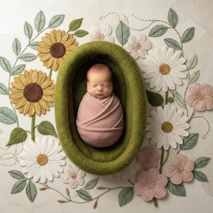 May include: A swaddled newborn baby in a green felt nest, surrounded by embroidered flowers. The flowers include sunflowers, daisies, and other blooms in yellow, white, and pink. The overall theme is floral and delicate.
