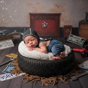May include: A newborn baby wearing a denim jumpsuit and a gray cap with white stitching, sleeping peacefully on a fluffy white cushion atop a black tire. The scene is set with vintage car parts and license plates.