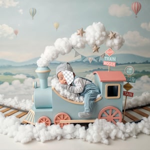 May include: A newborn baby wearing a gray hat and blue overalls is nestled in a light blue toy train. The train has pink accents and is surrounded by fluffy white clouds. Hot air balloons float in the background.