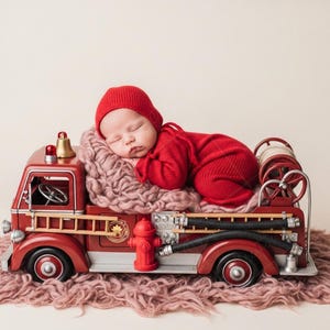 May include: A newborn baby wearing a red knit outfit and hat, sleeping peacefully on a pink blanket inside a vintage red fire truck prop. The fire truck has silver and black details.