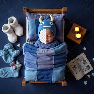 Puede incluir: Un bebé con un gorro y atuendo de monstruo azul, durmiendo en una cama de madera en miniatura con un edredón de retazos. La escena incluye calcetines blancos, calcetines azules, un libro y decoraciones en forma de estrella, todo sobre una superficie azul oscuro.