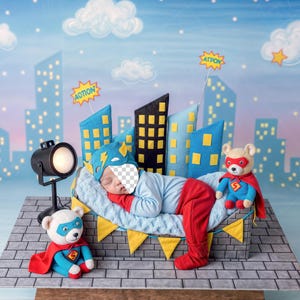 May include: A newborn baby dressed as a superhero, lying on a bed with a cityscape backdrop. The baby wears a blue and red costume with a matching hat. Two teddy bears in superhero outfits are also present.