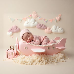 Könnte beinhalten: Ein Neugeborenes in rosa Schlafanzug und passendem Hut ruht in einer rosa Flugzeug-Requisite. Das Flugzeug sitzt auf einem Bett aus flauschigem weißem Material, mit einem kleinen rosa Koffer in der Nähe. Dekorative Wolken und ein Banner sind im Hintergrund.