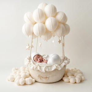 Pode incluir: Um recém-nascido dormindo em uma cesta branca em forma de balão de ar quente, cercado por pompons brancos. O balão é decorado com nuvens de tecido branco e estrelas douradas.