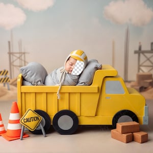 May include: A baby wearing a yellow construction hat, sleeping in a yellow toy dump truck. The scene includes toy construction cones, a caution sign, and toy bricks. The truck is surrounded by a construction site backdrop.