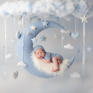 Puede incluir: Un gran accesorio de media luna azul claro texturizado con forro de piel sintética blanca, suspendido bajo una nube azul esponjosa. De la nube cuelgan varias estrellas, corazones y nubes de tela pequeña con cintas blancas, creando una escena celestial.